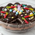 Easy Easter dirt cake layered with chocolate pudding, crushed cookies, and whipped topping, decorated with candy eggs and gummy worms, set in a cheerful springtime setting.