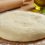 Soft and elastic homemade Italian pizza dough on a floured wooden surface, with a baker’s hands stretching it, surrounded by olive oil, basil, and a rustic kitchen backdrop