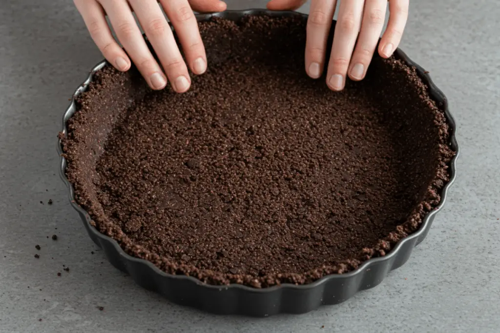 A dark chocolate cookie crust being pressed into a pie dish, forming a rich and crispy base for the Ravenclaw-inspired cheesecake.