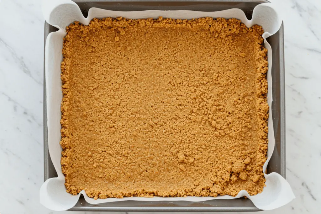 A buttery graham cracker crust being pressed into a baking pan, forming the base for the cheesecake bars.