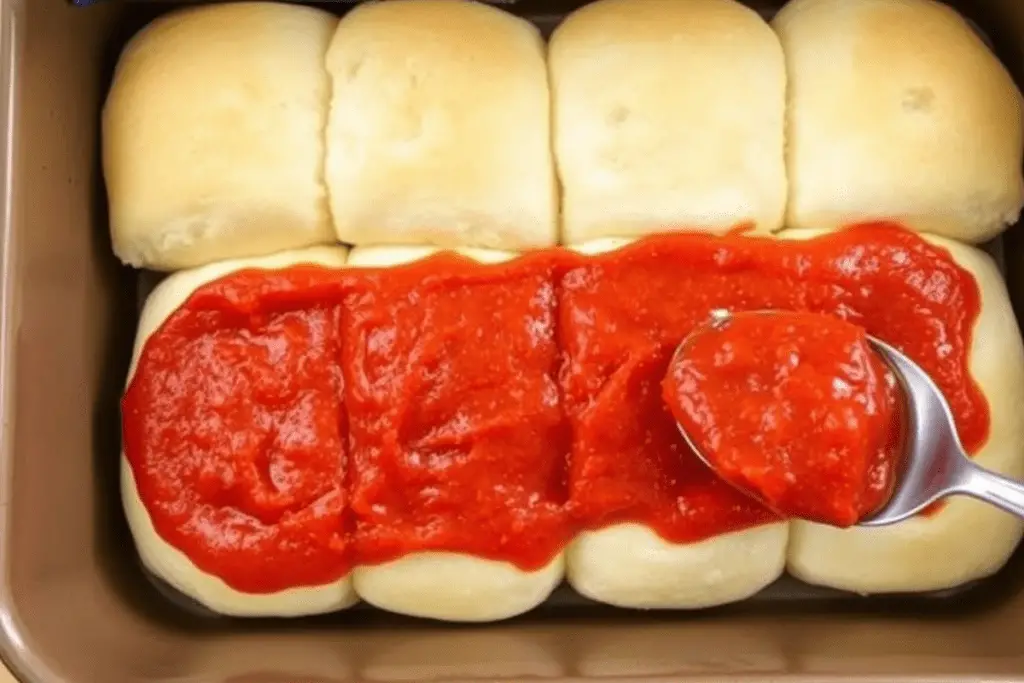 A baking dish with Hawaiian rolls sliced in half, topped with a layer of flavorful pizza sauce for Pepperoni Pizza Sliders.